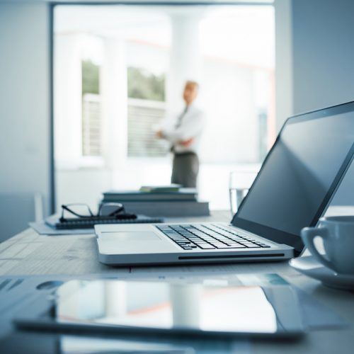 Businessman standing with arms crossed in his office, desktop with laptop on foreground, selective focus